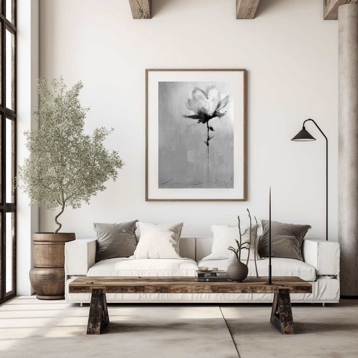 A monochrome painting of a single flower, rendered in shades of gray and black, hangs on a white wall above a white couch. The couch features gray and white pillows. A rustic wooden coffee table sits in front of the couch, and a small olive tree in a wooden pot is placed to the left. A floor lamp with a black shade stands in the corner. The overall aesthetic is minimalist and modern.