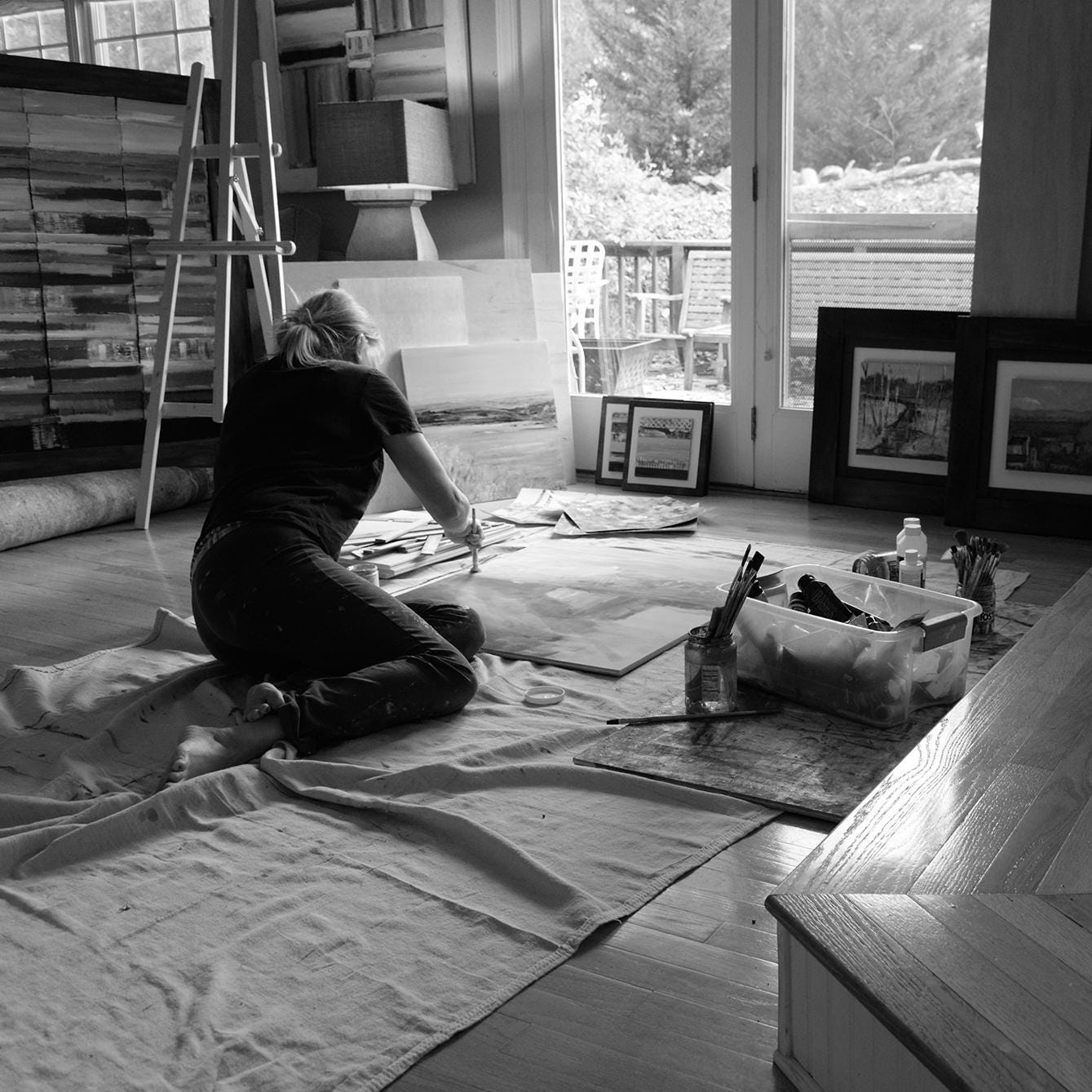 Artist Sallie Otenasek at work in studio with finished pieces in the background. Sallie is sitting on the floor, she prefers to work that way rather than standing at an easel.