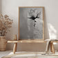 A monochromatic framed canvas print featuring a stylized flower in shades of gray. The flower is depicted with loose brushstrokes, creating a textured and painterly effect. The background is a soft, muted gray, providing a subtle contrast to the darker tones of the flower. The overall style is minimalist and modern, with a focus on the artistic expression of the floral subject. The print is displayed on a light-colored wooden bench, adding to the serene and calming atmosphere of the scene.