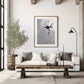 A monochrome painting of a single flower, rendered in shades of gray and black, hangs on a white wall above a white couch. The couch features gray and white pillows. A rustic wooden coffee table sits in front of the couch, and a small olive tree in a wooden pot is placed to the left. A floor lamp with a black shade stands in the corner. The overall aesthetic is minimalist and modern.
