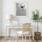A grayscale abstract floral painting of a single flower hangs above a white wooden desk.  A laptop, lamp, and chair are on the desk. A small olive tree in a woven basket sits on the floor next to the desk. The overall aesthetic is minimalist and modern.