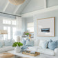 May include: A white couch with blue pillows in a living room with a large framed painting of a blue boat on a white background. The painting is hanging on a light blue wall. The couch is in front of a large window with white curtains. There is a coffee table in front of the couch with a plant and a basket of books on it.