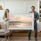 May include: A framed landscape painting of a lake with a pink and blue sky. The painting is in a white frame and is being held by two people.