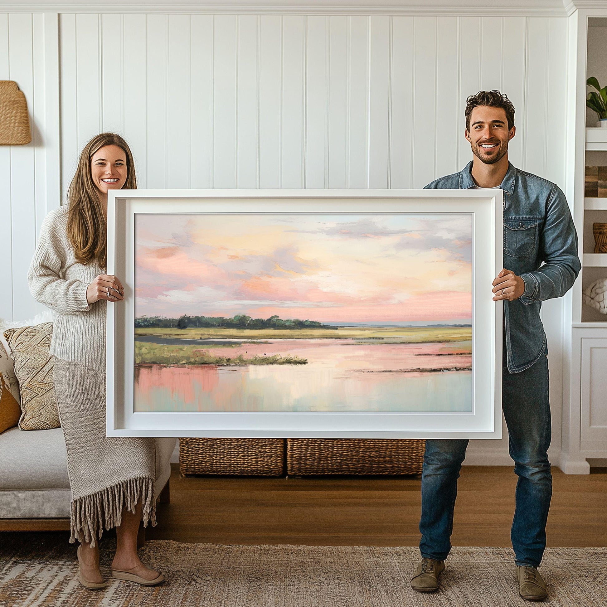 May include: A framed landscape painting of a lake with a pink and blue sky. The painting is in a white frame and is being held by two people.