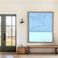 May include: An abstract painting with a light blue upper section and a lighter blue lower section, framed in a natural wood. The artwork hangs above a woven bench with wooden legs, placed on a neutral-toned rug. A woven basket sits near a dark green door with glass panels, and a black sconce is mounted on the wall.