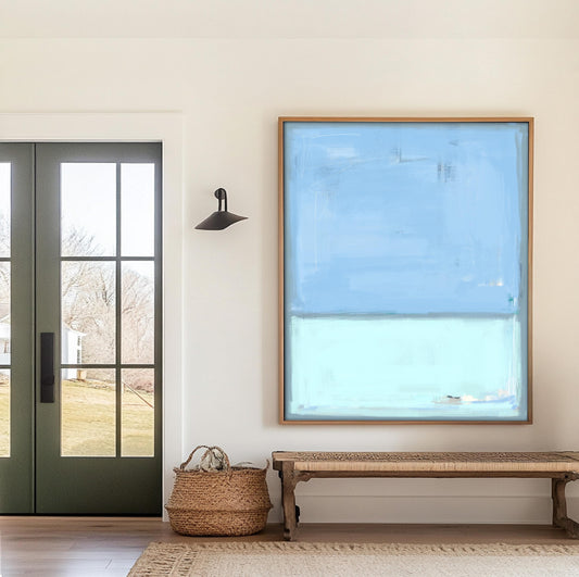May include: An abstract painting with a light blue upper section and a lighter blue lower section, framed in a natural wood. The artwork hangs above a woven bench with wooden legs, placed on a neutral-toned rug. A woven basket sits near a dark green door with glass panels, and a black sconce is mounted on the wall.