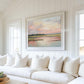 May include: A white couch with white pillows in front of a large framed painting of a pink and blue sunset over a body of water.