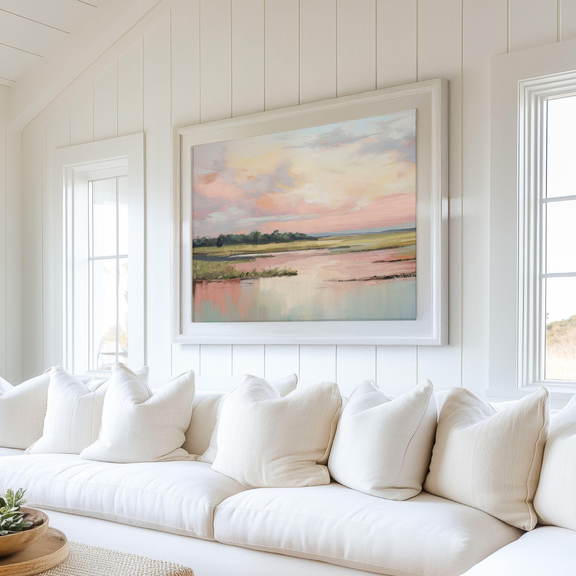 May include: A white couch with white pillows in front of a large framed painting of a pink and blue sunset over a body of water.