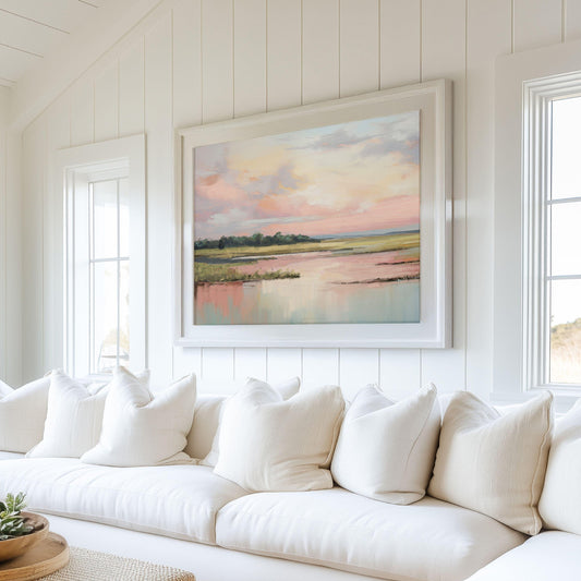 May include: A white couch with white pillows in front of a large framed painting of a pink and blue sunset over a body of water.