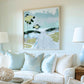 May include: An abstract painting in a silver frame hangs above a white sofa with light blue pillows. The artwork features yellow and black floral shapes and a road in shades of blue, green, and white. Two matching lamps flank the sofa.