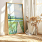May include: A framed landscape painting with a gold frame. The artwork depicts a serene scene of a wooden pier leading to a body of water, surrounded by green grass and trees. The sky is a soft blue with hints of clouds.